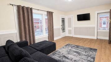 A living room with a sofa set, a patterned rug, a flat-screen TV, and a door leading to a balcony.