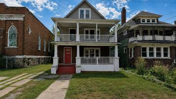 A two-story house with a front porch, a red door, and a brick building to the left.