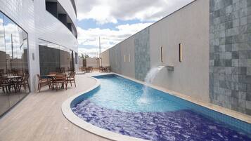 Una piscina interior con diseño curvo, rodeada de mesas y sillas de madera, y un edificio moderno con grandes ventanales y un muro de azulejos.