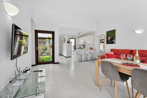 A modern living room with a dining area, a kitchen in the background, and a glass door leading to a garden. - Bungalow Island Village (Costa Adeje)