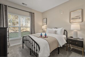 A hotel room with a bed, bedside tables, a window with blinds, a painting on the wall, and a view of trees outside. - New Albany 2BR Near Intel Easton Pets OK (Westerville)