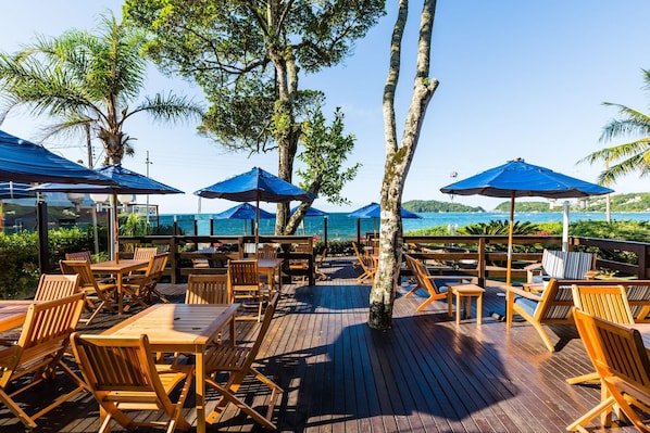 Un área de estar al aire libre con mesas y sillas de madera, sombrillas azules y vista al mar.