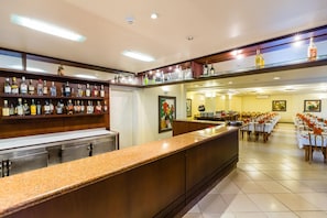 Un bar de hotel con una barra larga, estantes con botellas de licor y un área de comedor con mesas y sillas.