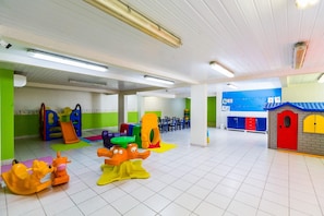 Un área de juegos interior bien iluminada con diversas estructuras de juego coloridas, una zona de estar con sillas y una pared azul con carteles educativos.