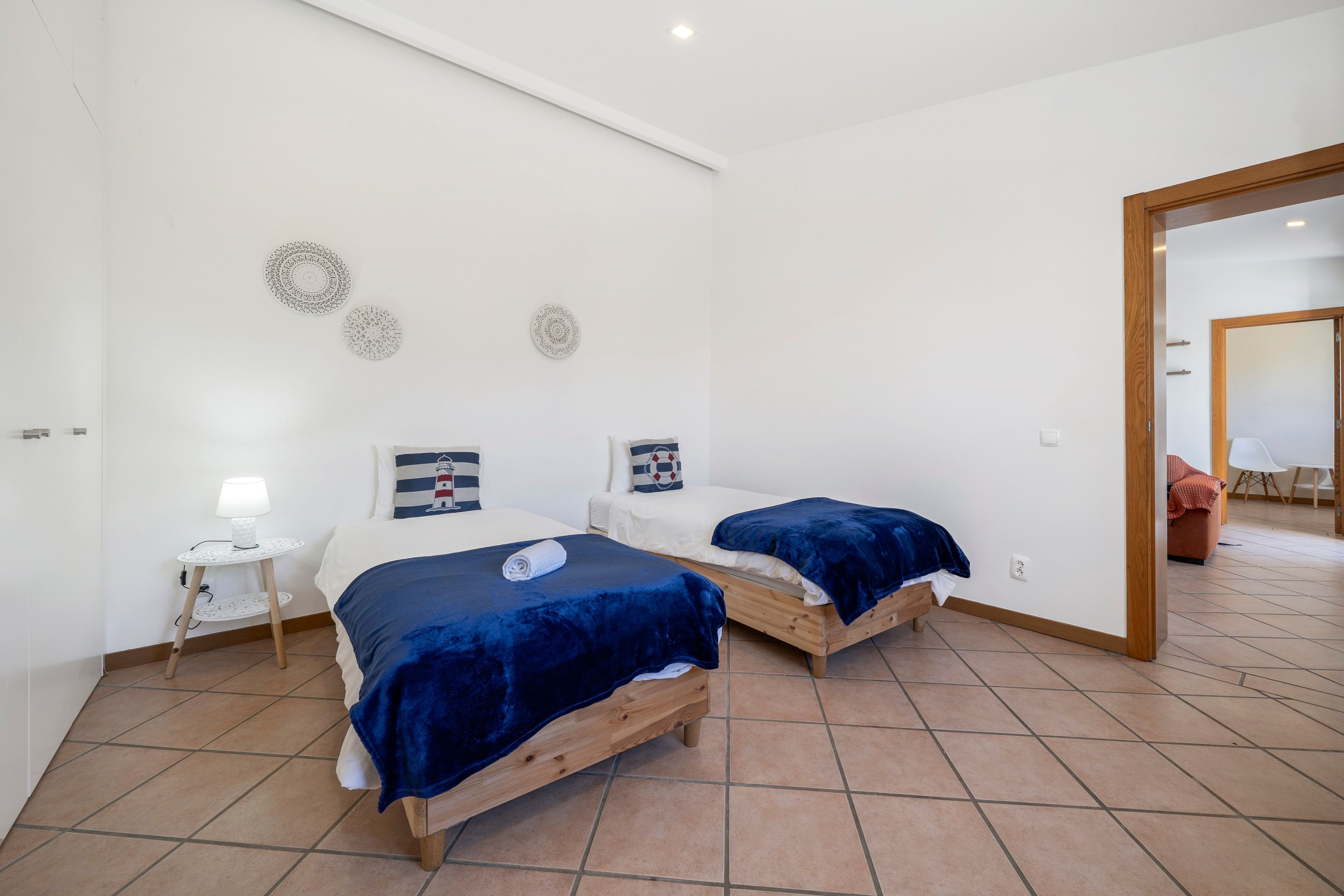 A bedroom with two beds, a small bedside table with a lamp, and decorative wall hangings.