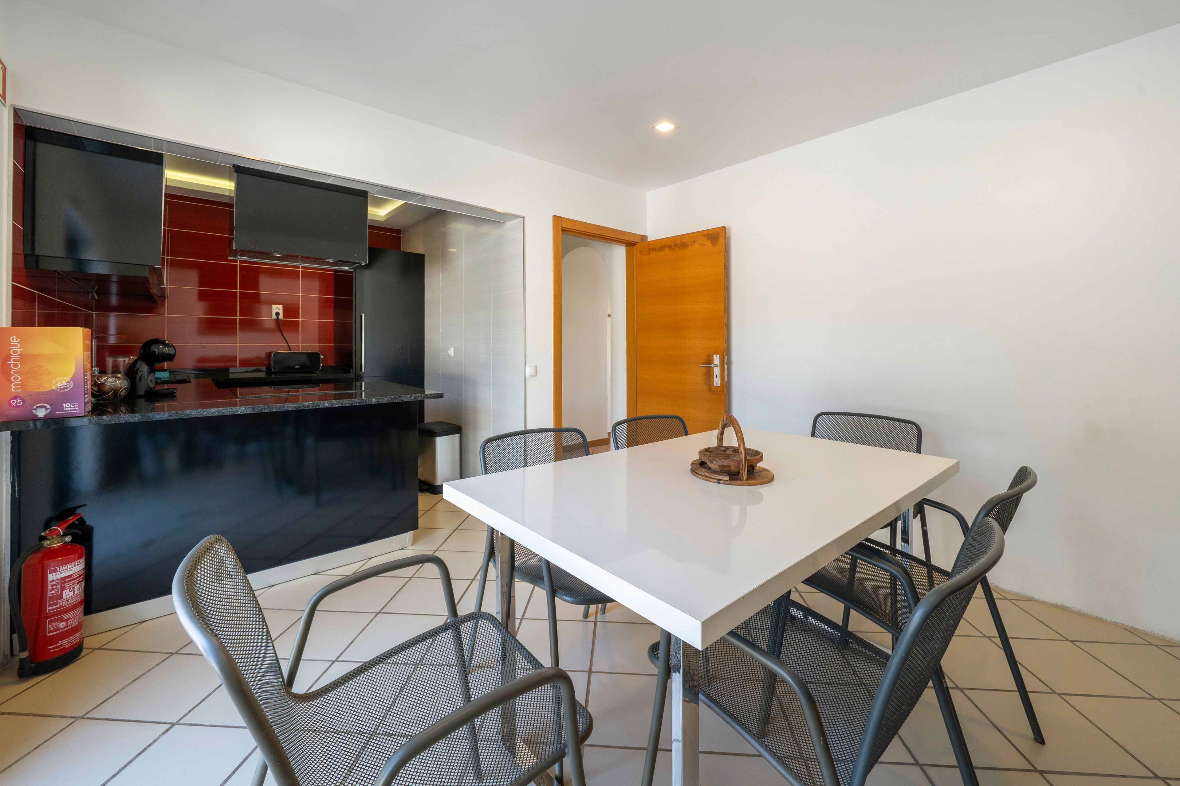 A modern kitchen with a white countertop, black cabinets, and a red backsplash. There is a dining table with six chairs and a wooden door in the background.