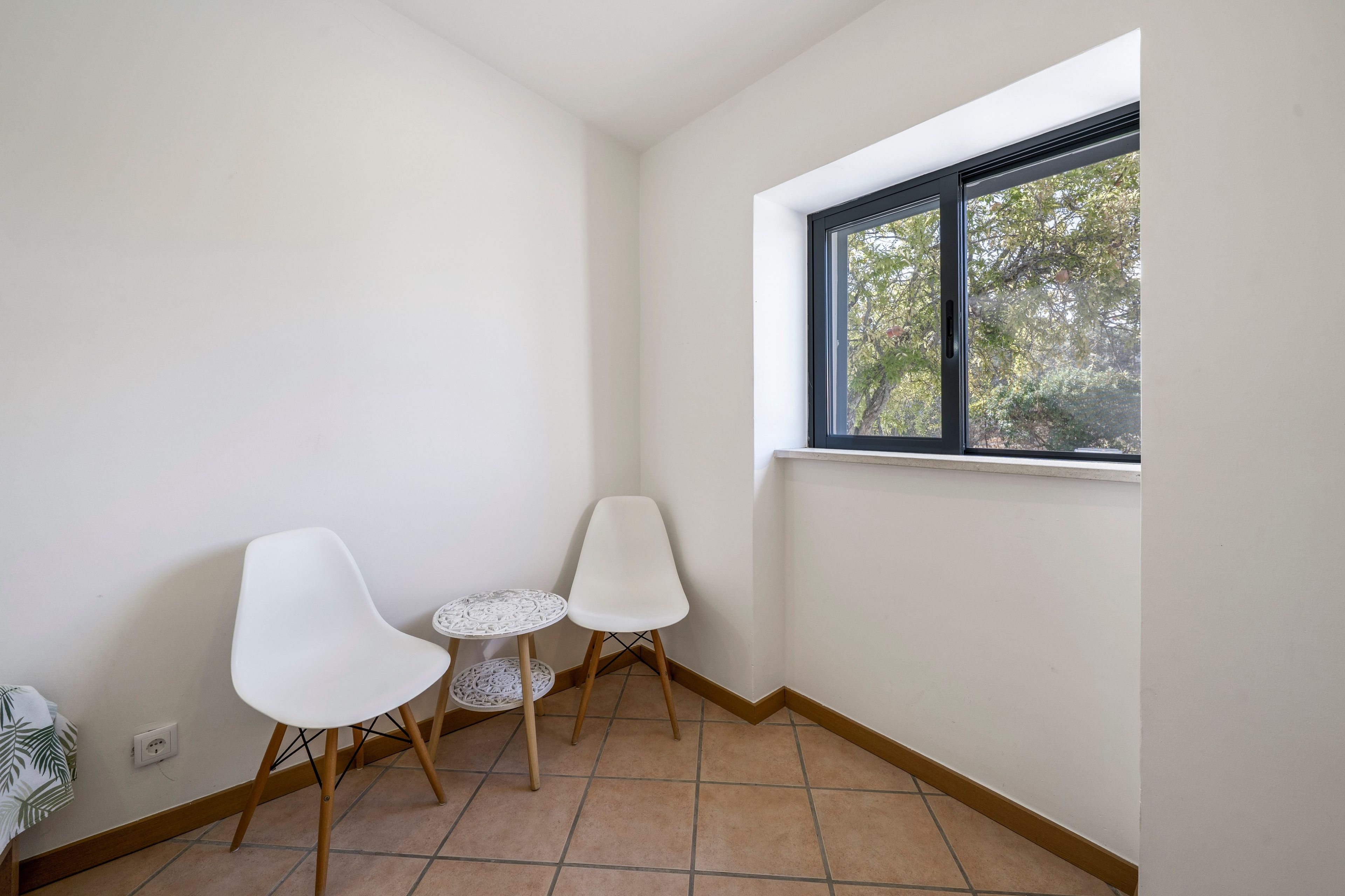A corner room with two white chairs, a small round table, and a window with a view of trees.
