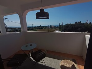 A balcony with a table and chairs, a hanging light fixture, and a view of a landscape with trees and buildings.