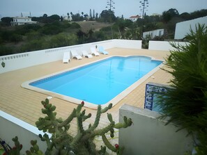 Une piscine dotée d’une terrasse en bois, entourée de verdure et avec un bâtiment blanc en arrière-plan.