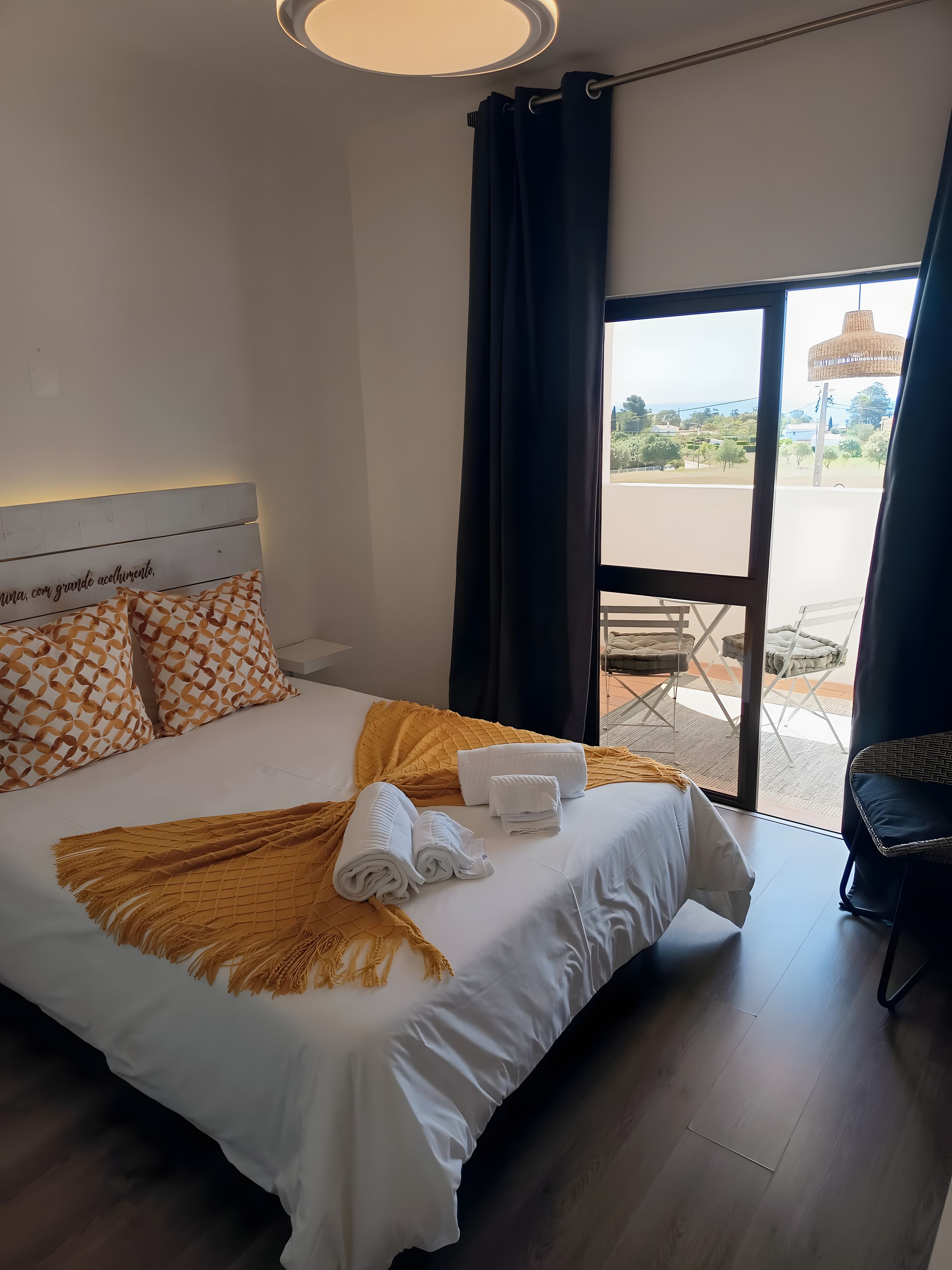Une chambre d’hôtel avec un lit, du linge de lit blanc, un plaid jaune et une vue sur l’extérieur grâce à une grande fenêtre.