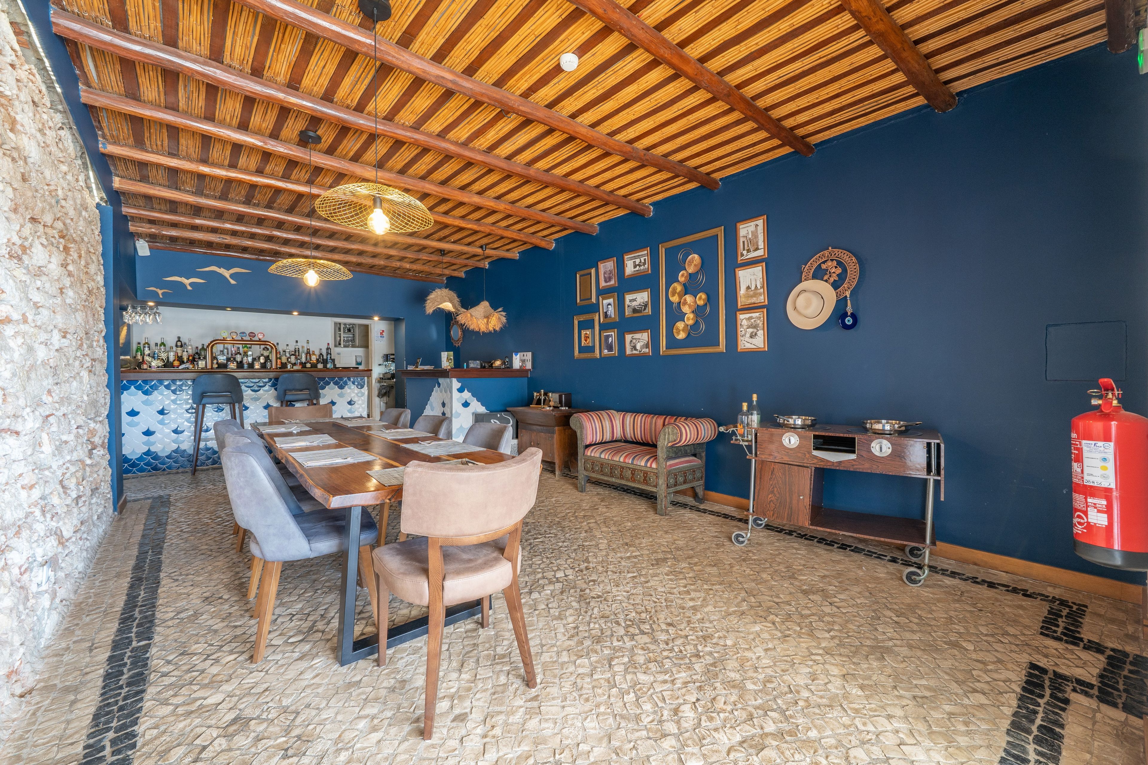 Een restaurant met een hoog houten plafond, een blauwe wand met ingelijste foto’s, een lange houten tafel met stoelen, en een brandblusser aan de rechterkant.