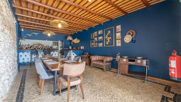 Een restaurant met een hoog houten plafond, een blauwe wand met ingelijste foto’s, een lange houten tafel met stoelen, en een brandblusser aan de rechterkant.