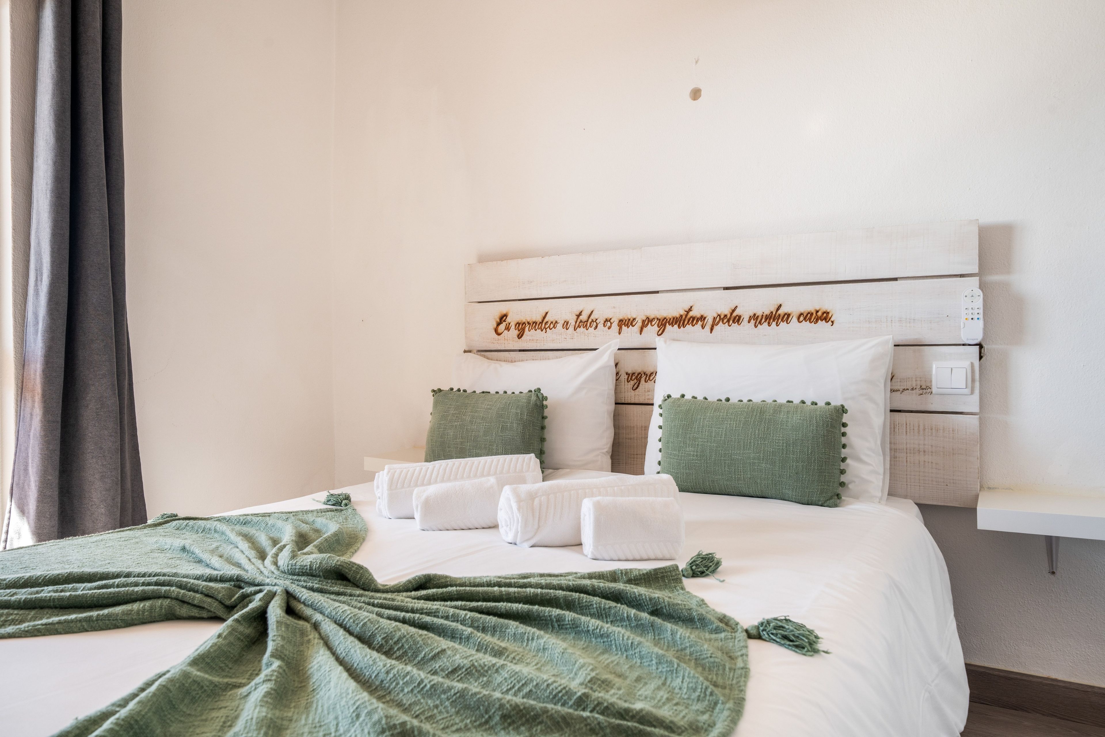 Een net opgemaakt bed met witte lakens en een groen gebreide deken, kussens met groene kwastjes, en een houten hoofdbord met een decoratief plaquette.