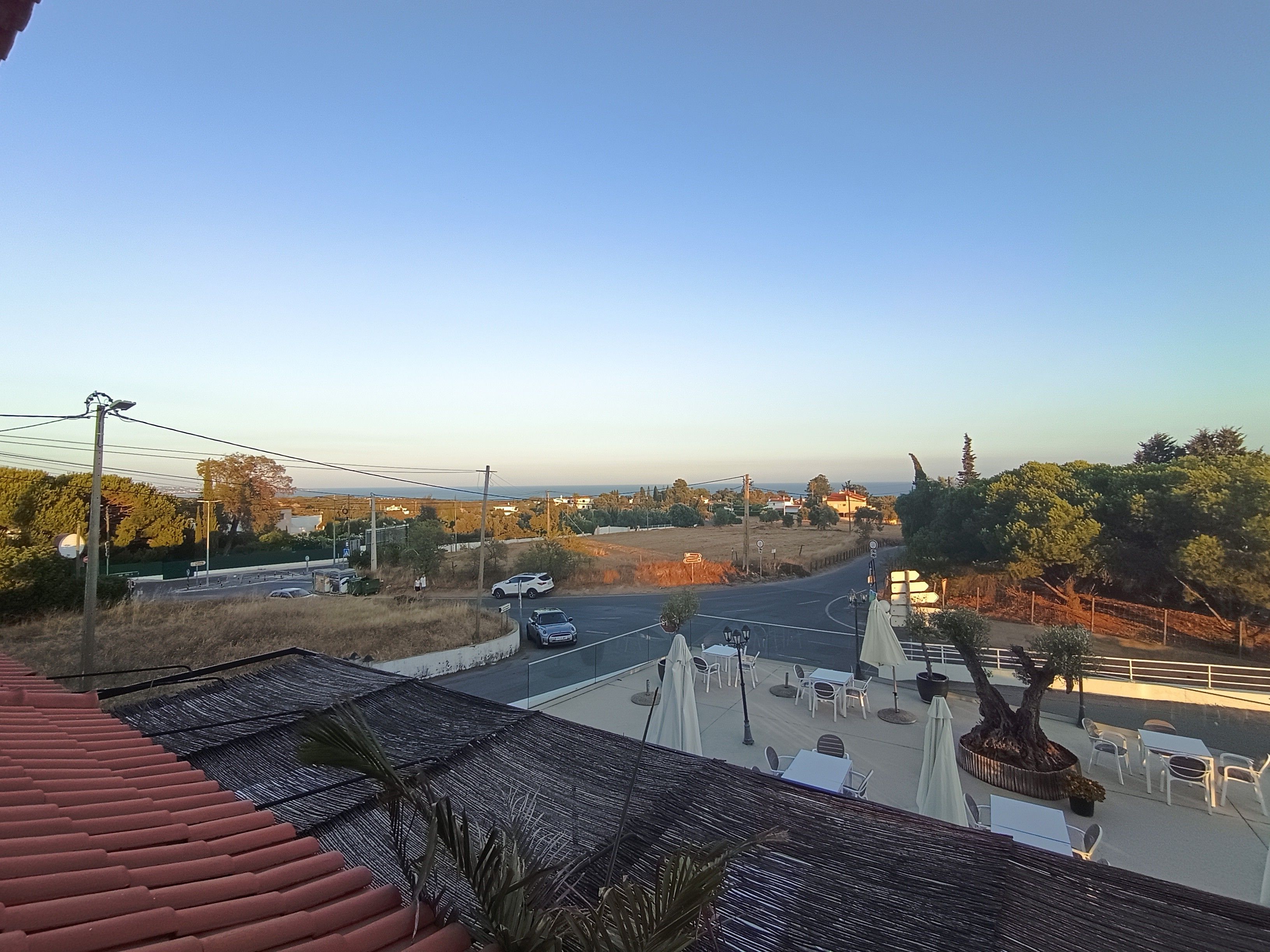 Vista de uma estrada com veículos, uma faixa de pedestres e um edifício com mesas ao ar livre.