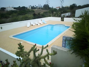 A swimming pool with a wooden deck, surrounded by greenery and a white building in the background.