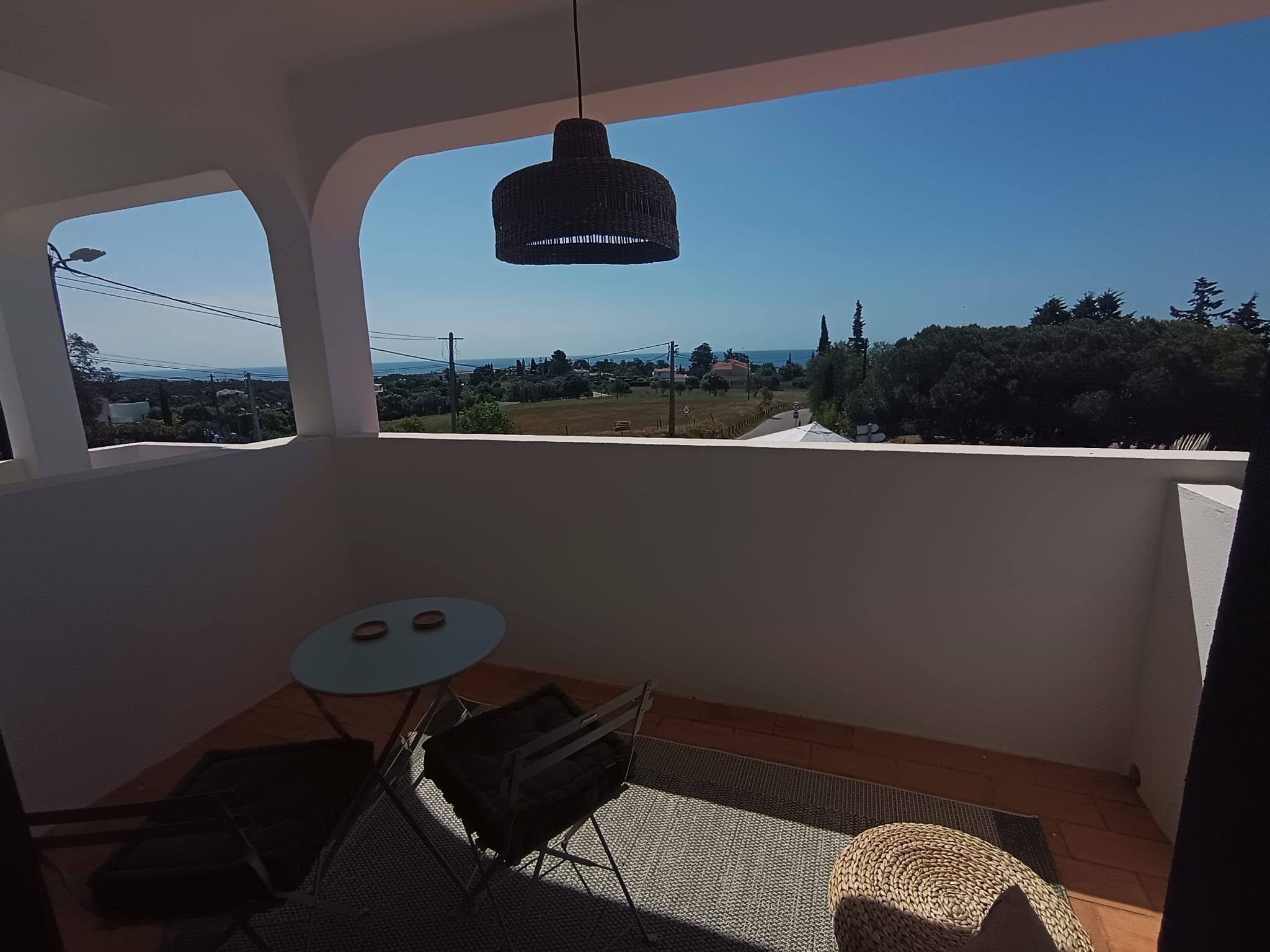 Ein Balkon mit Tisch und Stühlen, einer Hängelampe und Blick auf eine Landschaft mit Bäumen und Gebäuden.