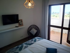 A bedroom with a bed, a television on the wall, a chair, and a large window.