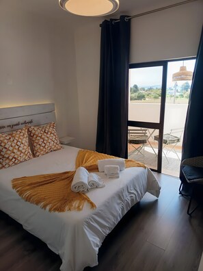 Ein Hotelzimmer mit einem Bett, weißer Bettwäsche, einer gelben Tagesdecke und Blick ins Freie durch ein großes Fenster.