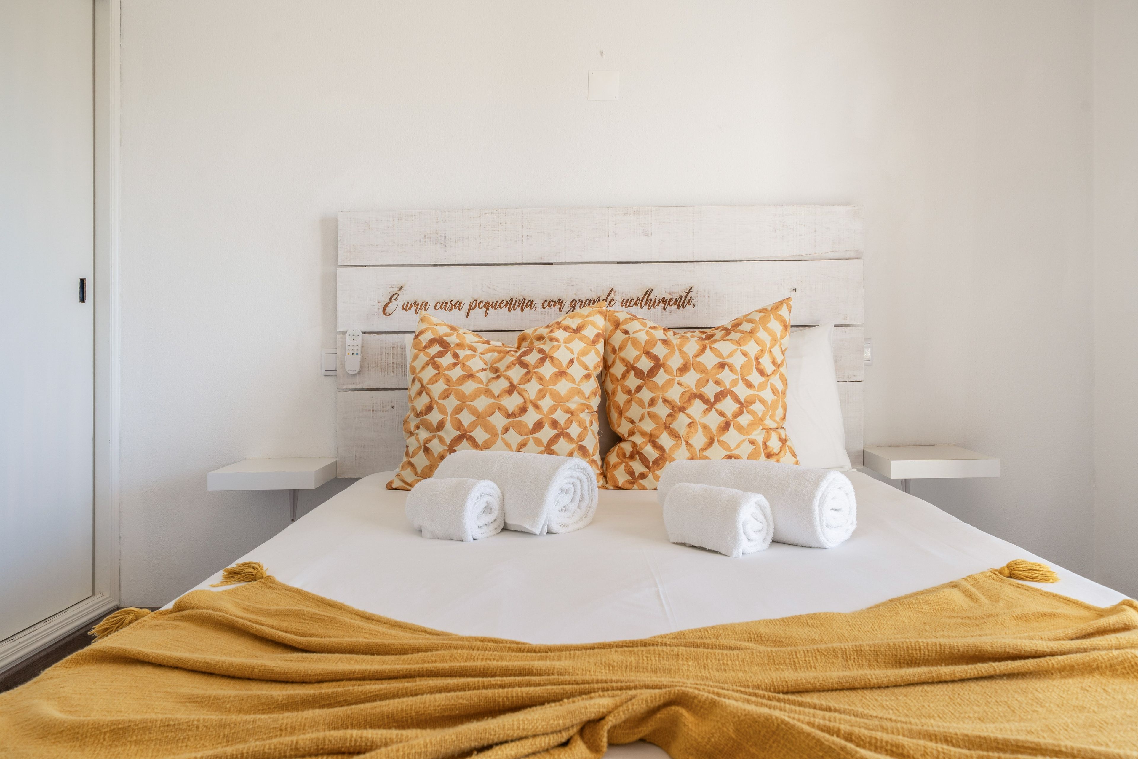Un lit bien fait, avec des oreillers et des serviettes enroulées, une tête de lit blanche ornée d’une inscription décorative et un plaid de couleur moutarde.