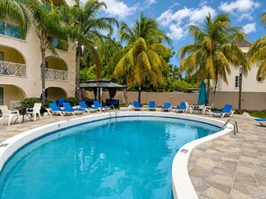 Ein Swimmingpool mit Liegestühlen und Sonnenschirmen, umgeben von Palmen und einem Gebäude mit Bögen.