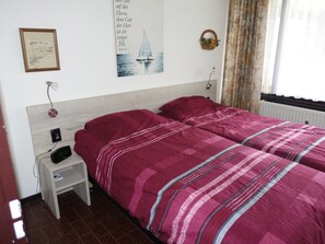 A bedroom with a bed, a nightstand, a lamp, a wall clock, a framed picture, and a window with curtains. - Holiday Apartment With 2 Bedrooms (De Koog)