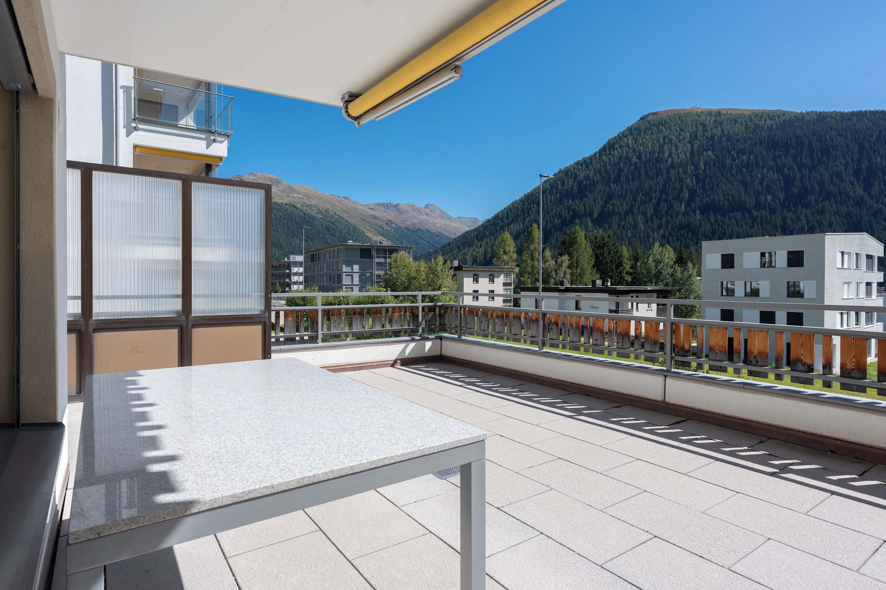 A balcony with a table and chairs, overlooking a mountainous landscape with buildings in the background.