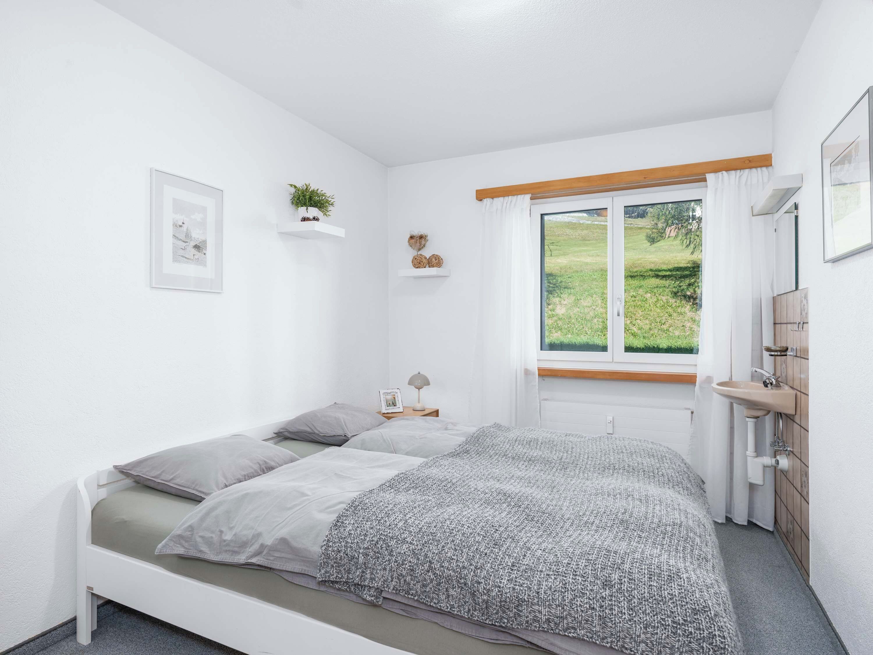 Ein Schlafzimmer mit einem Bett, einem Nachttisch, einer Lampe und einem Fenster mit Blick ins Grüne.