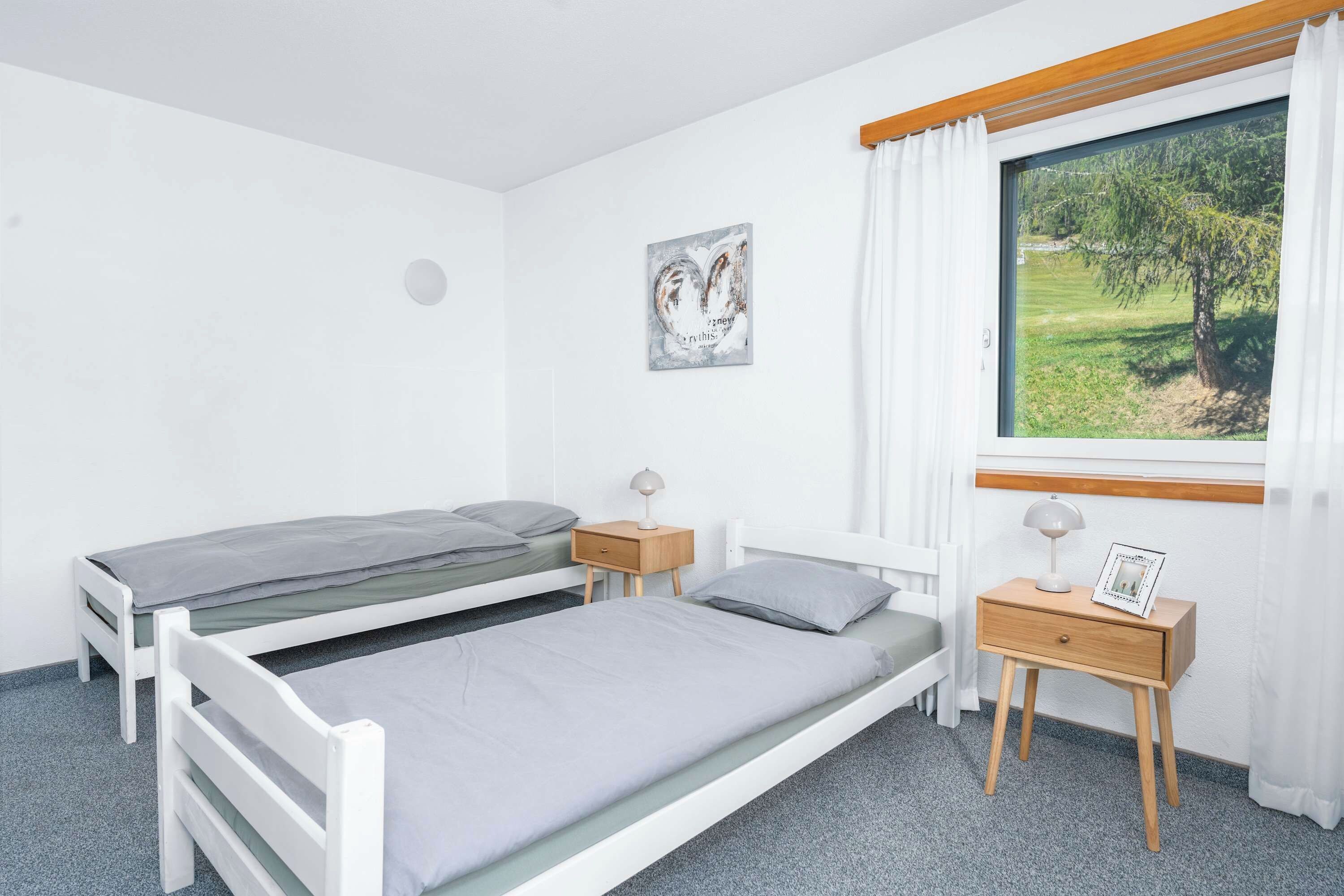 A bedroom with two single beds, a bedside table with a lamp, a small bedside table with a picture frame, a window with a view of greenery, and a wall-mounted artwork.