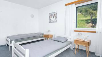 A bedroom with two single beds, a bedside table with a lamp, a small bedside table with a picture frame, a window with a view of greenery, and a wall-mounted artwork.