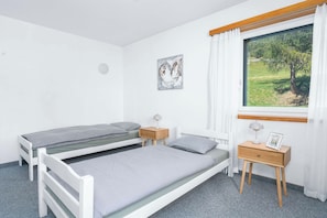 Ein Schlafzimmer mit zwei Einzelbetten, einem Nachttisch mit Lampe, einem kleinen Nachttisch mit Bilderrahmen, einem Fenster mit Blick auf Grünflächen und einem an der Wand hängenden Kunstwerk.