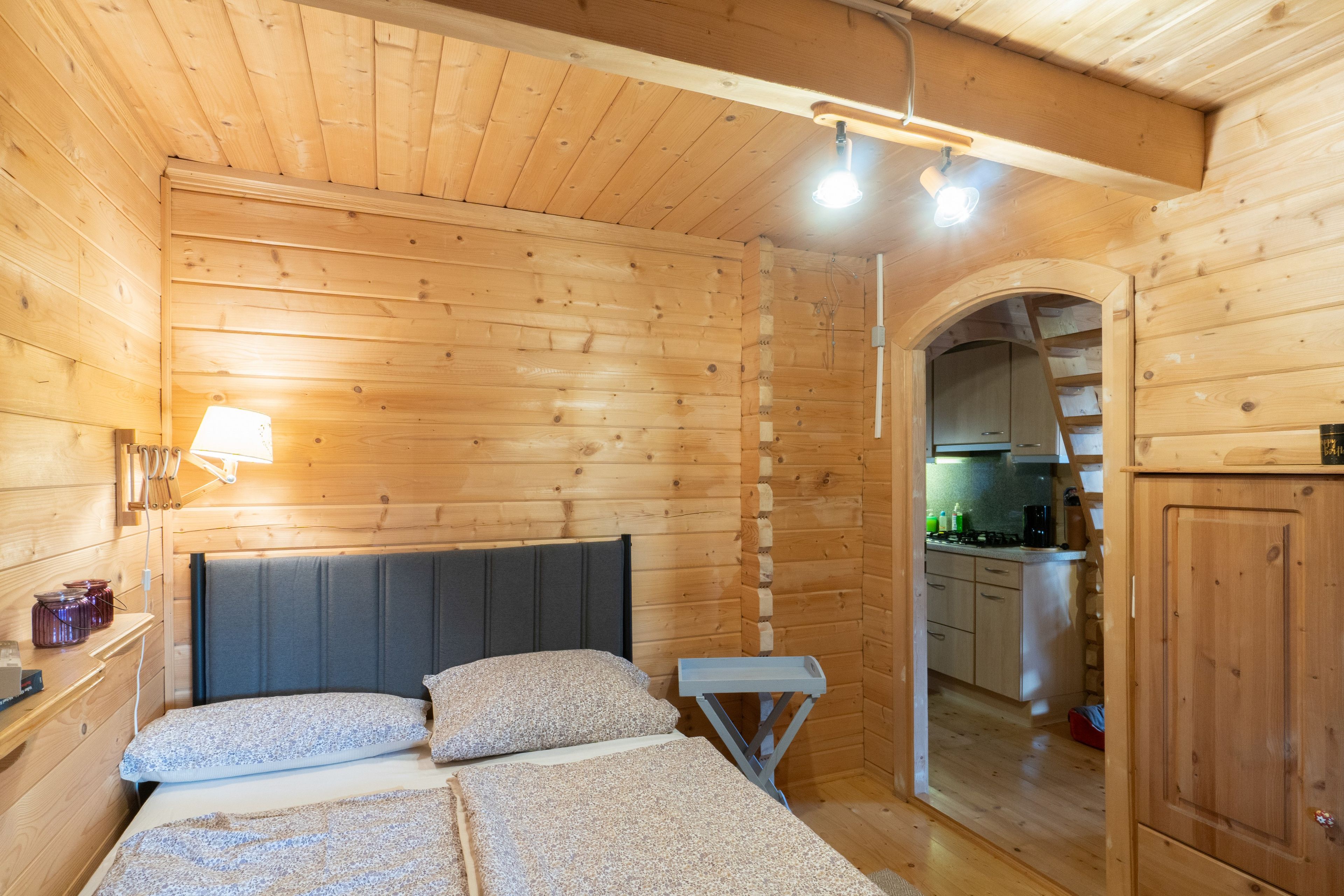 Ein Holzinnenraum mit einem Bett, einem Nachttisch, einer Lampe und Blick in einen Küchenbereich.