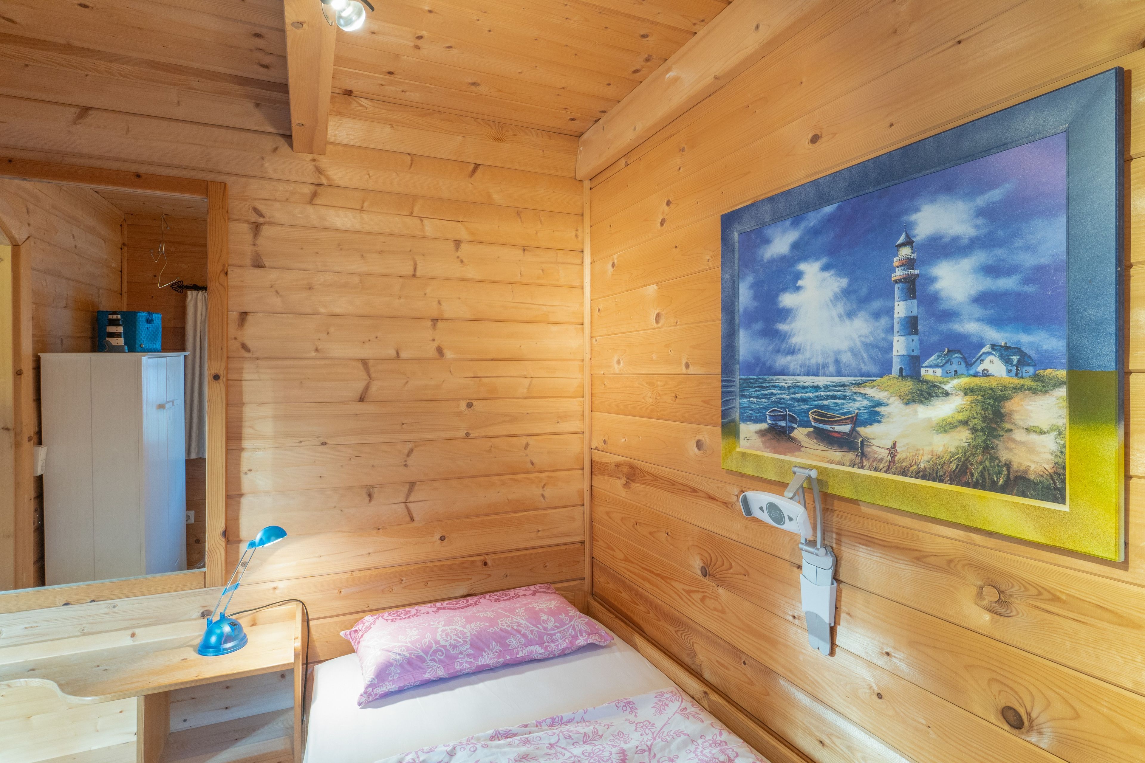 A wooden cabin room with a bed, a nightstand, a painting of a lighthouse, and a small shelf.