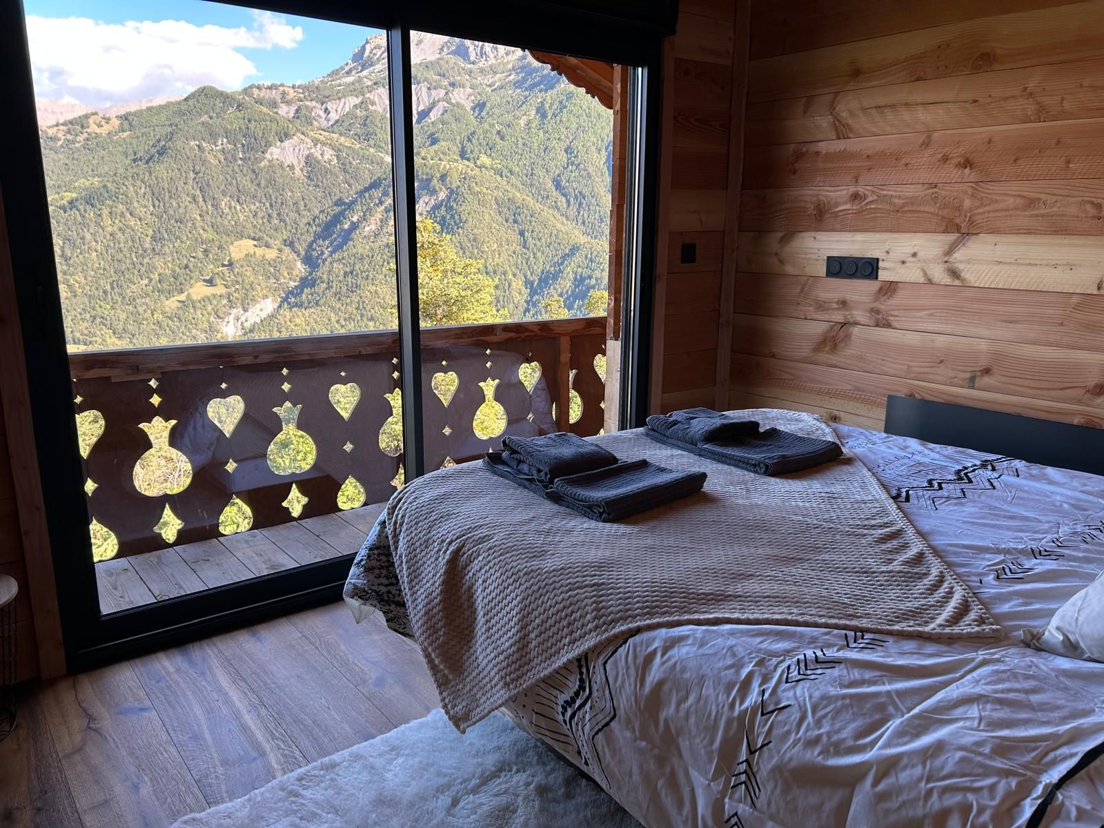 Un dormitorio con una ventana grande, paredes de madera, una cama con colcha estampada y vistas a las montañas.