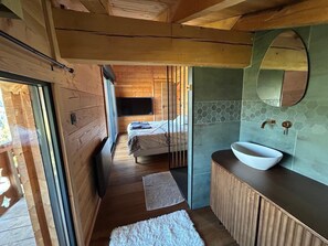 Een houten interieur met een bed, een badkamer met een grote spiegel en wastafel, en uitzicht door een glazen deur.
