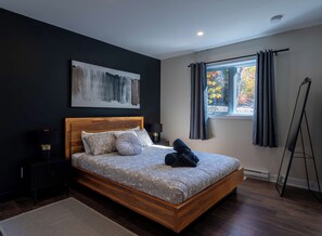 A bedroom with a wooden bed, a painting on the wall, a window with curtains, and a floor lamp. - L’Eden | Modern chalet with spa, foosball table, and 2 living rooms (Saint-Côme)