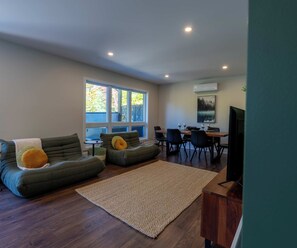 A modern living room with a sofa set, a dining table with chairs, a TV, and a large window with a view of trees. - L’Eden | Modern chalet with spa, foosball table, and 2 living rooms (Saint-Côme)