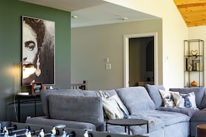 A living room with a grey sectional sofa, a coffee table, a wall-mounted shelf with decorative items, and a large abstract painting. - L’Eden | Modern chalet with spa, foosball table, and 2 living rooms (Saint-Côme)