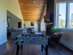 A cozy living room with a foosball table, a fireplace, and a large window offering a view of trees. - L’Eden | Modern chalet with spa, foosball table, and 2 living rooms (Saint-Côme)