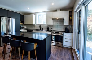 A modern kitchen with a central island, bar stools, stainless steel appliances, and a view through a large window. - L’Eden | Modern chalet with spa, foosball table, and 2 living rooms (Saint-Côme)