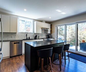 A modern kitchen with a central island, bar stools, and a large window. - L’Eden | Modern chalet with spa, foosball table, and 2 living rooms (Saint-Côme)