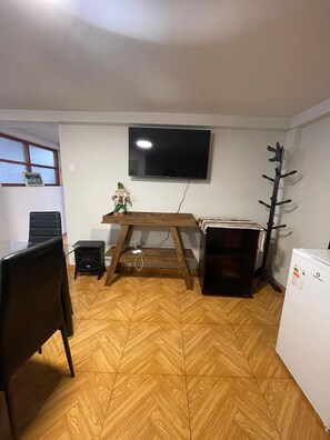 Ein Zimmer mit Holzdielen im Fischgrätmuster, einem an der Wand montierten Fernseher, einem kleinen Tisch mit einer dekorativen Figur, einem Kleiderständer und einem Kühlschrank.