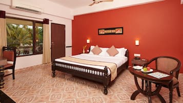 A hotel room with a bed, a chair, a table, a window with curtains, a wall-mounted air conditioner, and a ceiling fan.