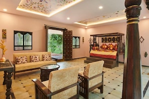 A room with a sofa, armchairs, a bed with a colorful bedspread, a decorative ceiling, and a view of greenery outside. - Sterling Palavelli Godavari (Elamanchili)