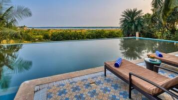 An infinity pool with lounge chairs and a tiled deck, overlooking a landscape with greenery and a distant view.