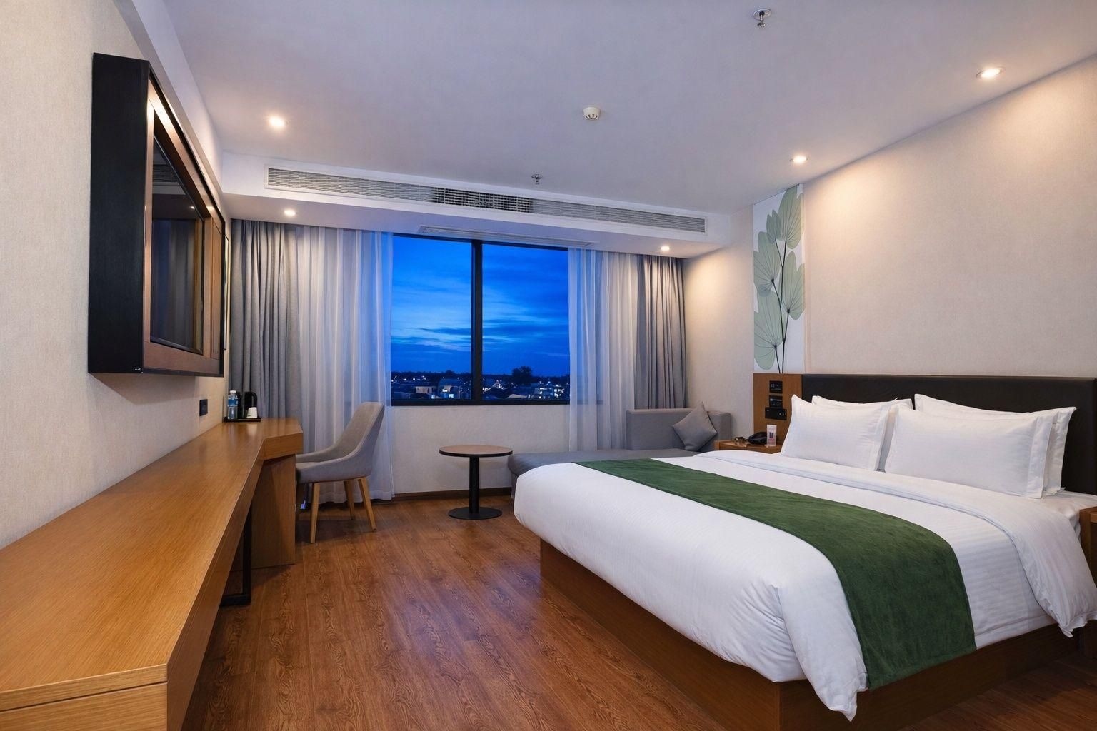 Quarto de hotel com cama, escrivaninha, cadeira, mesinha e uma grande janela com vista para a cidade ao entardecer.