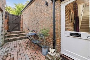 A brick exterior with a white door, a small outdoor table with chairs, and a metal trash bin. - The Barn A Cosy Countryside Retreat Near Eastbourne (Eastbourne)
