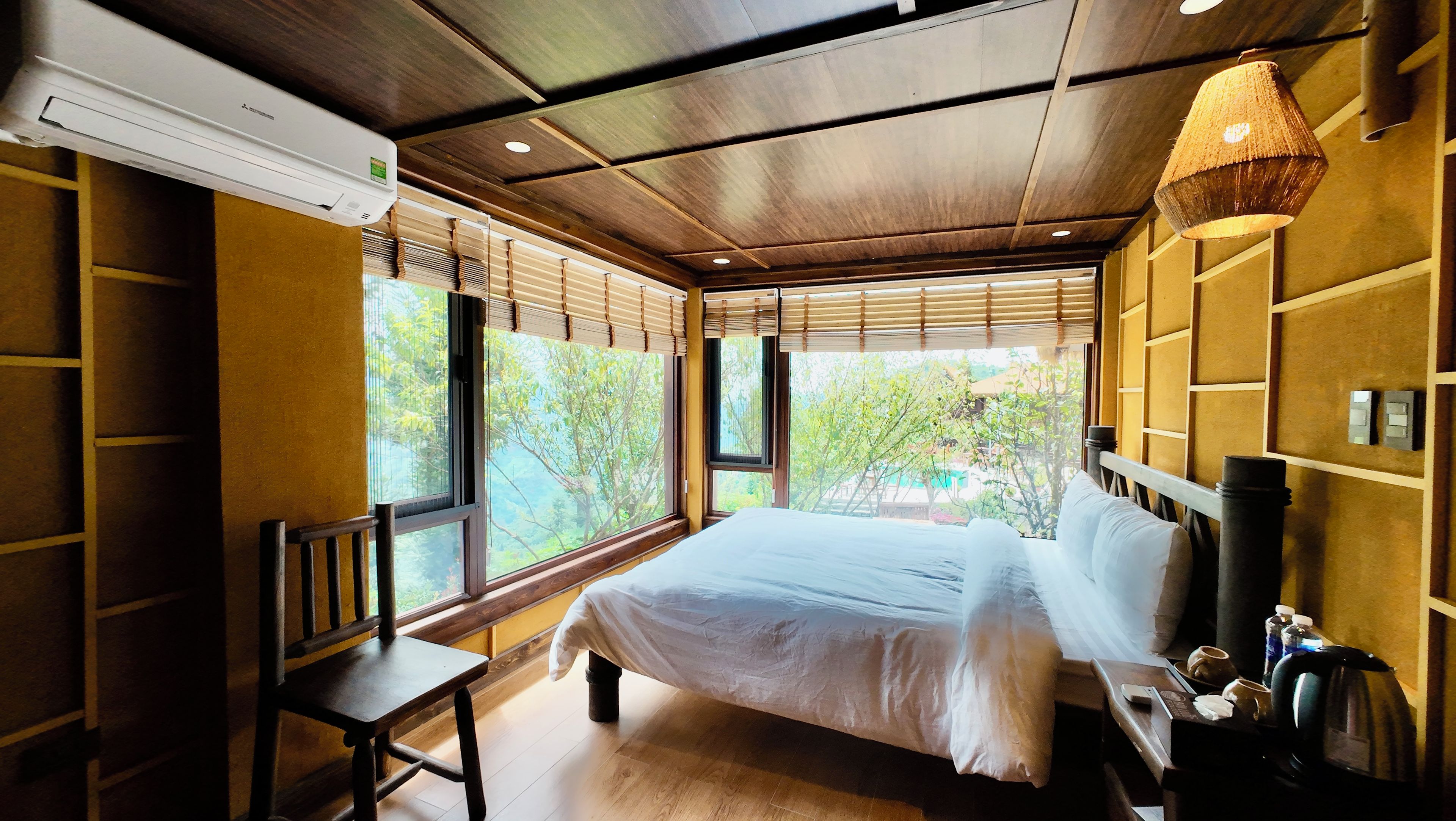 Quarto com cama, mesa de cabeceira e janela com vista para o verde.