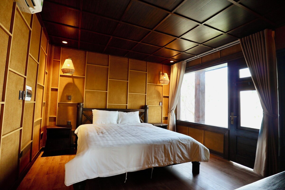 A hotel room with a wooden ceiling, a bed with white bedding, two bedside lamps, a nightstand, and a window with curtains.