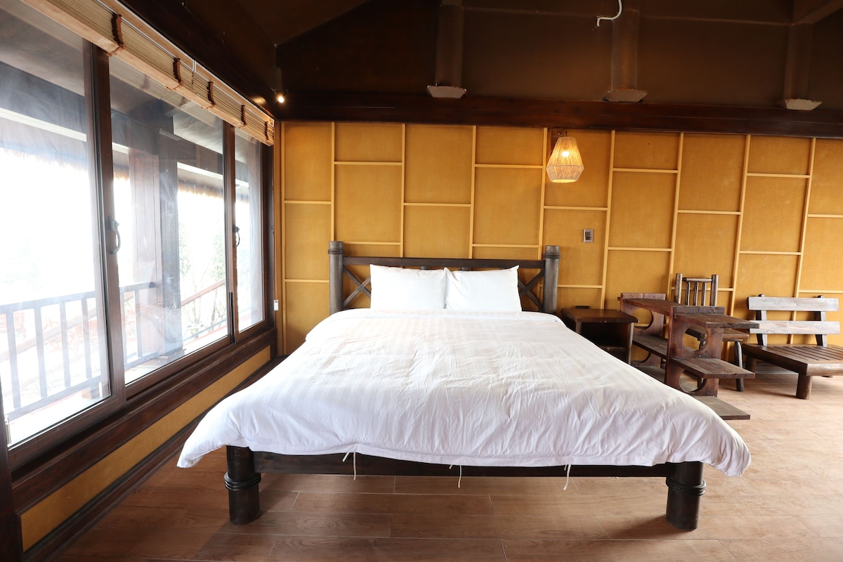 Una cama grande con cabecera de madera, suelo de madera, ventana con vistas y una mesa con sillas.
