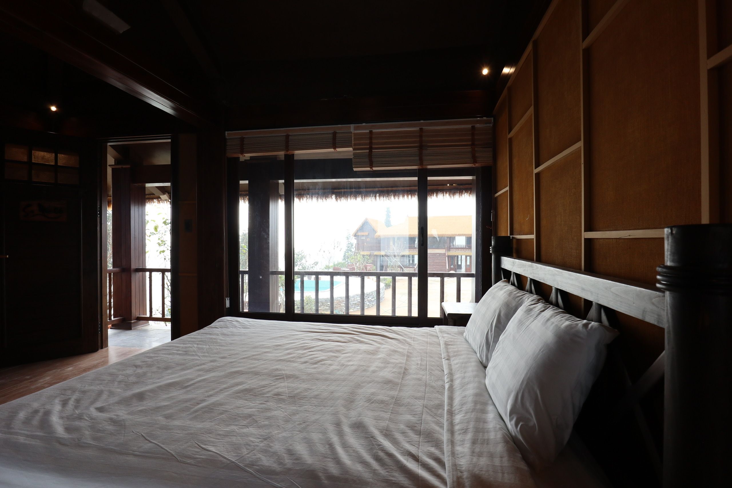 A large bed with a wooden headboard, a view of a building through a window, and a wooden floor.